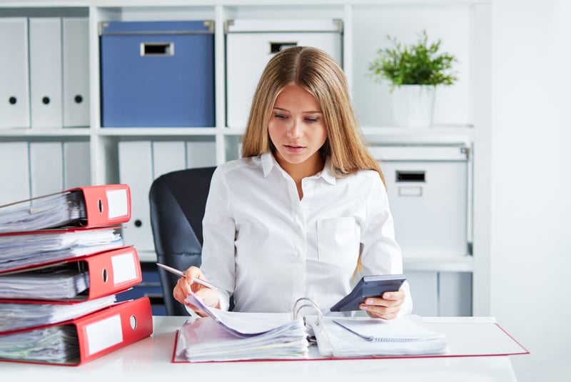 A Woman Sitting at a Desk Holding a Calculator and Looking in a Ring Binder